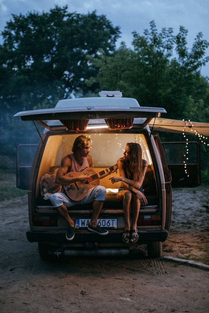 Relaxing couple playing guitar inside camper van during outdoor evening.
